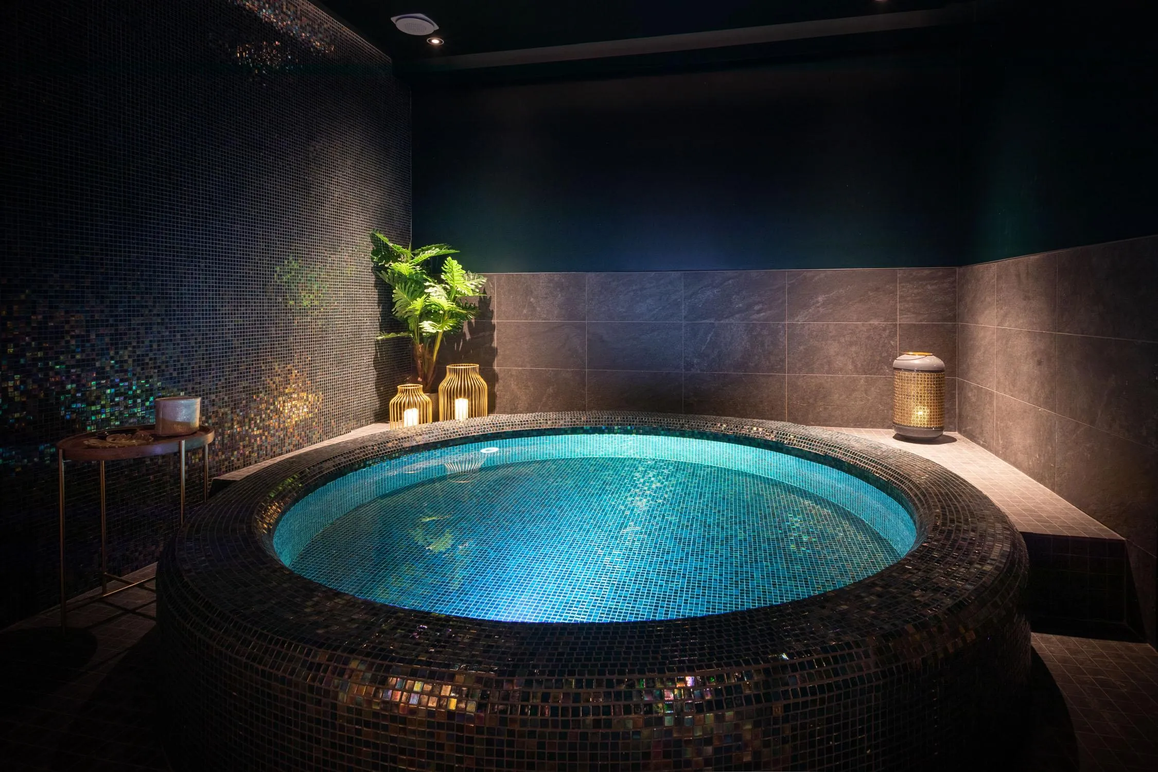 Jacuzzi circulaire avec éclairage bleu entouré de mosaïques, dans une pièce aux murs sombres avec des plantes et des lanternes