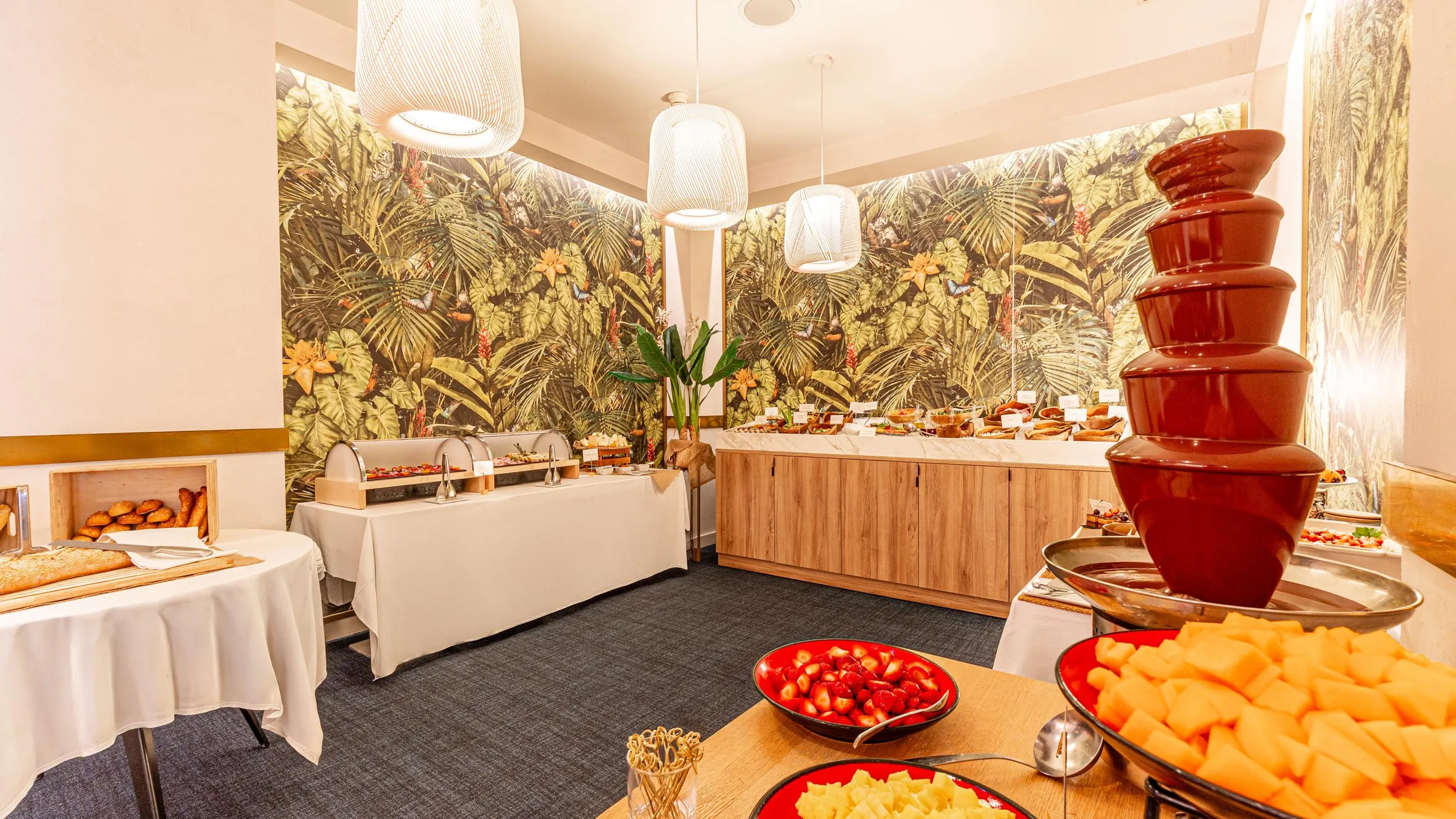 Buffet avec divers plats, dont une fontaine à chocolat, dans une salle décorée de motifs tropicaux et éclairée par des suspensions