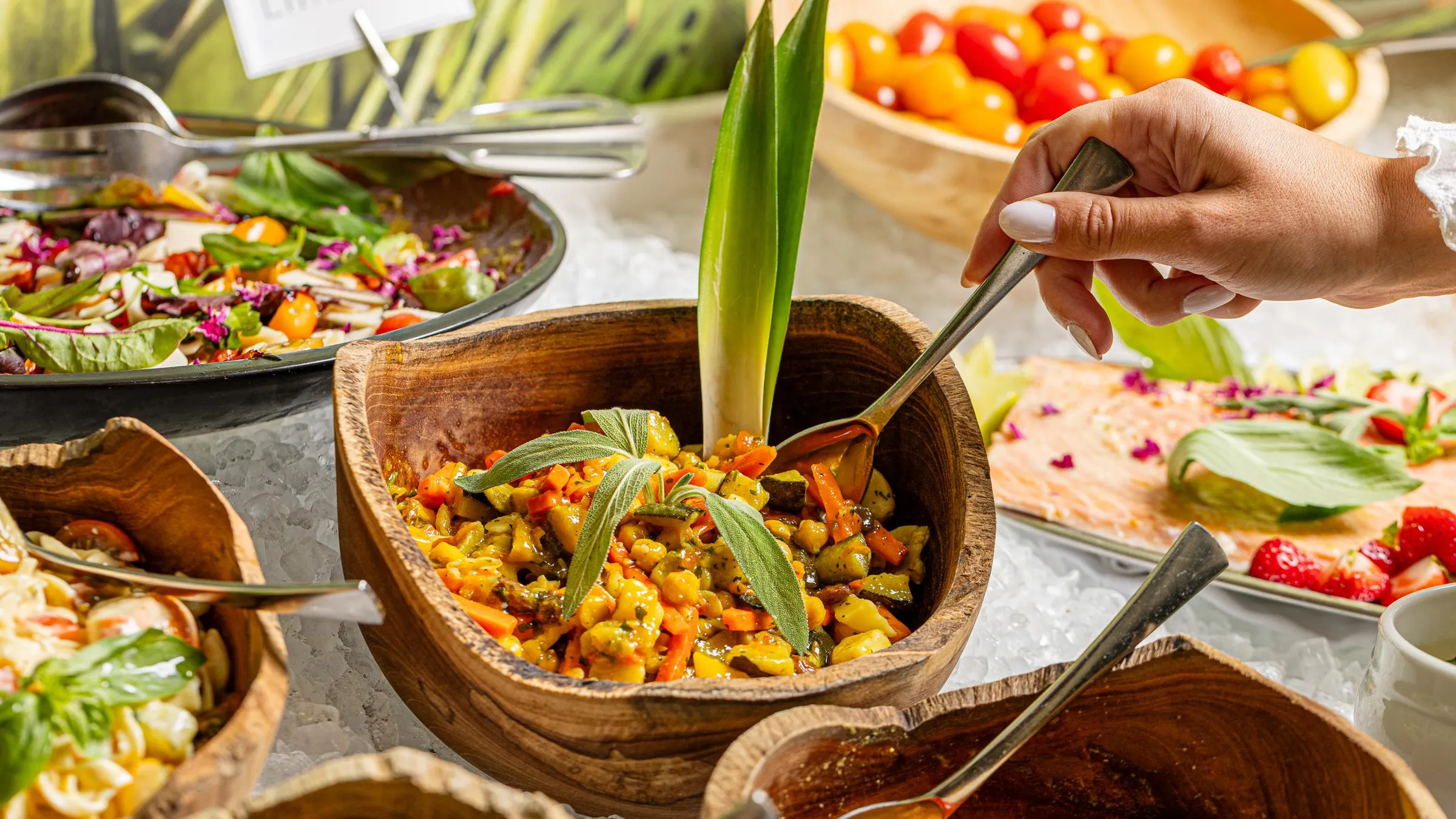 Une main sert une préparation de légumes dans un bol en bois sur un buffet de plats variés