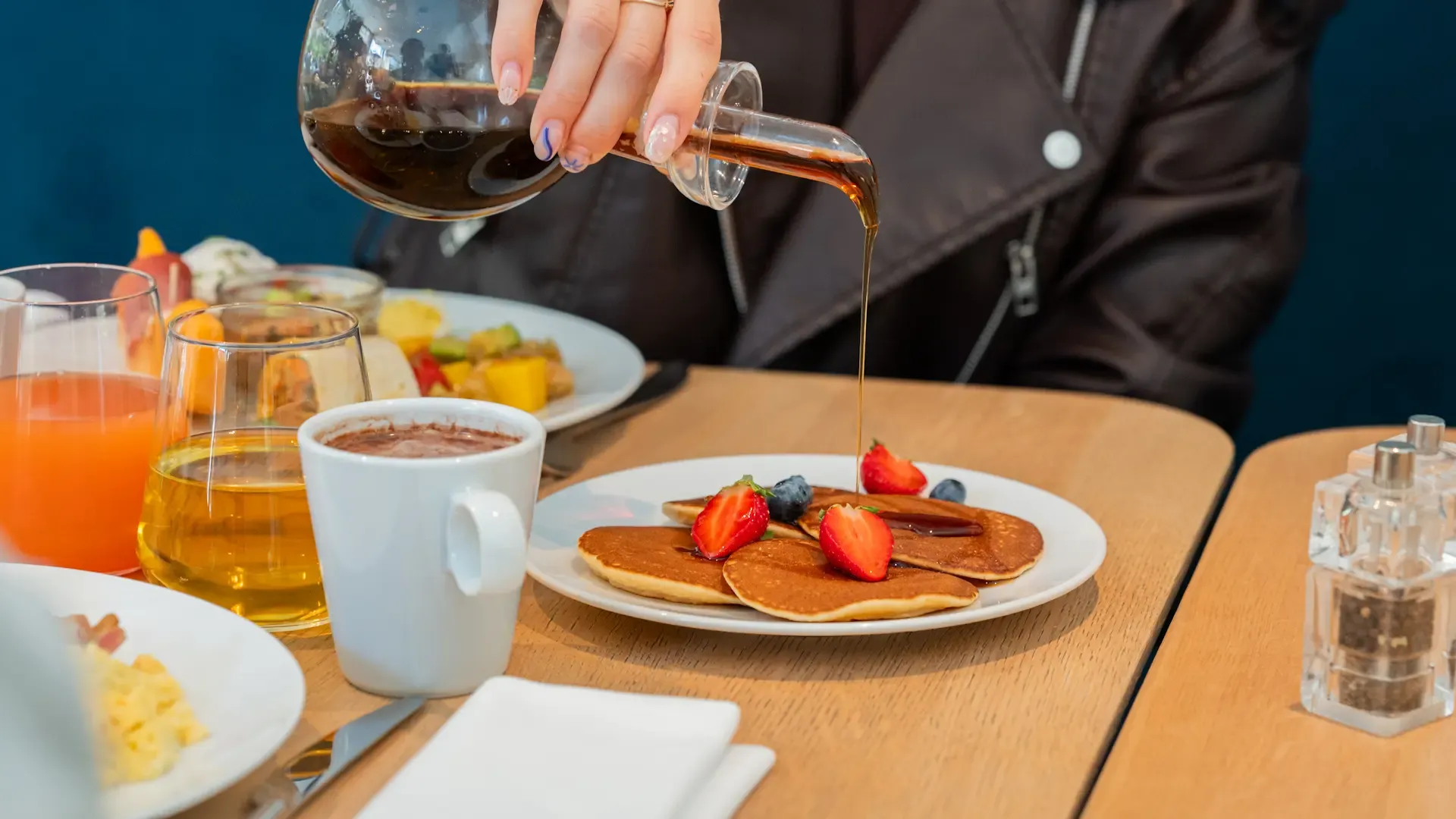 Une personne verse du sirop sur des pancakes garnis de fruits, accompagnés de boissons et d'un plateau de fruits sur une table