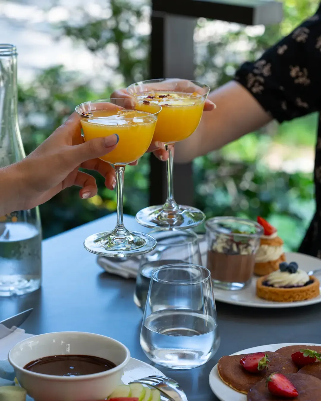 Deux mains lèvent des verres à cocktail orange garnis de zestes, autour d'une table avec des desserts et des boissons