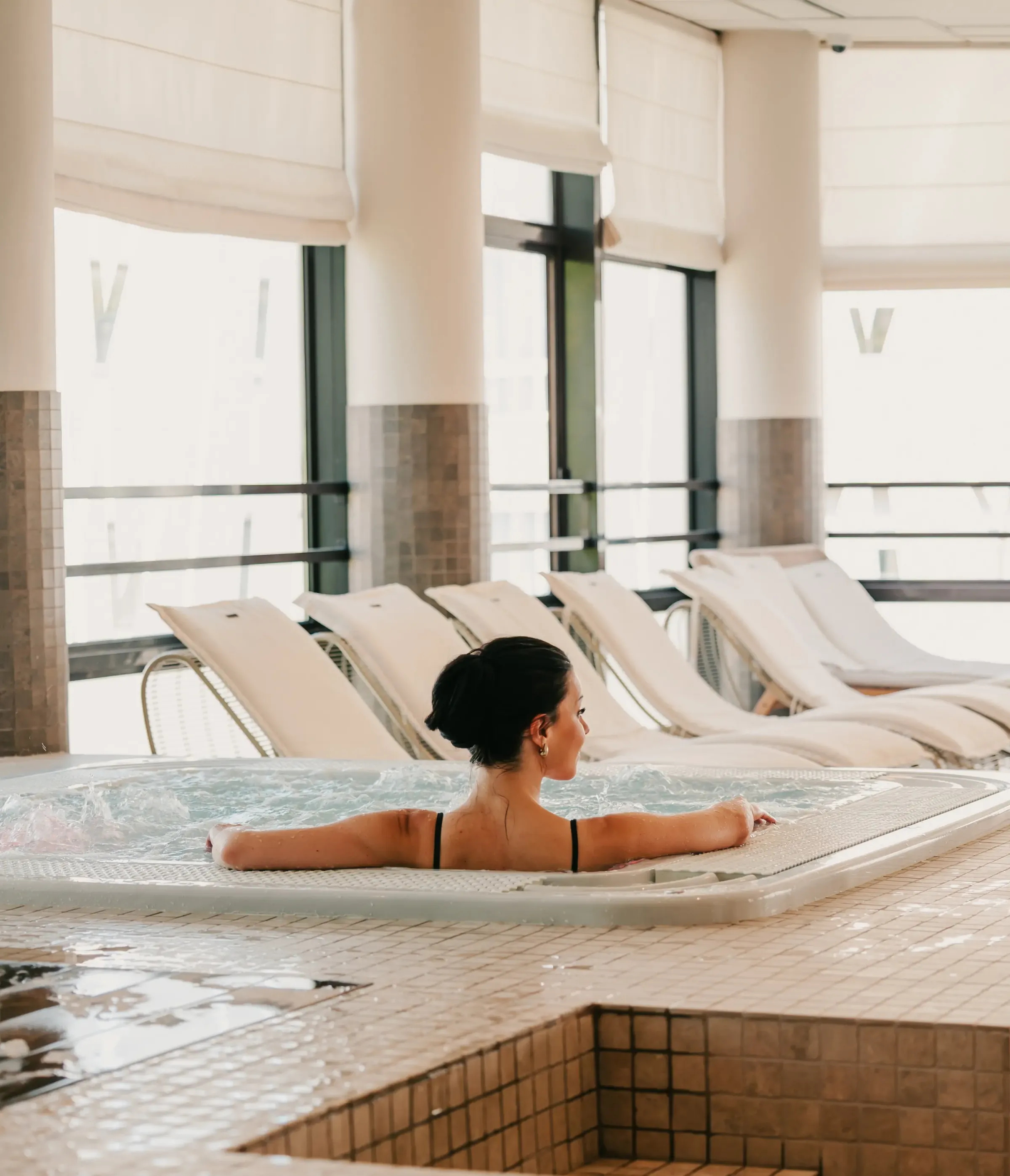 Une femme en maillot de bain noir est allongée dans un bain à remous, entourée de transats près de grandes fenêtres
