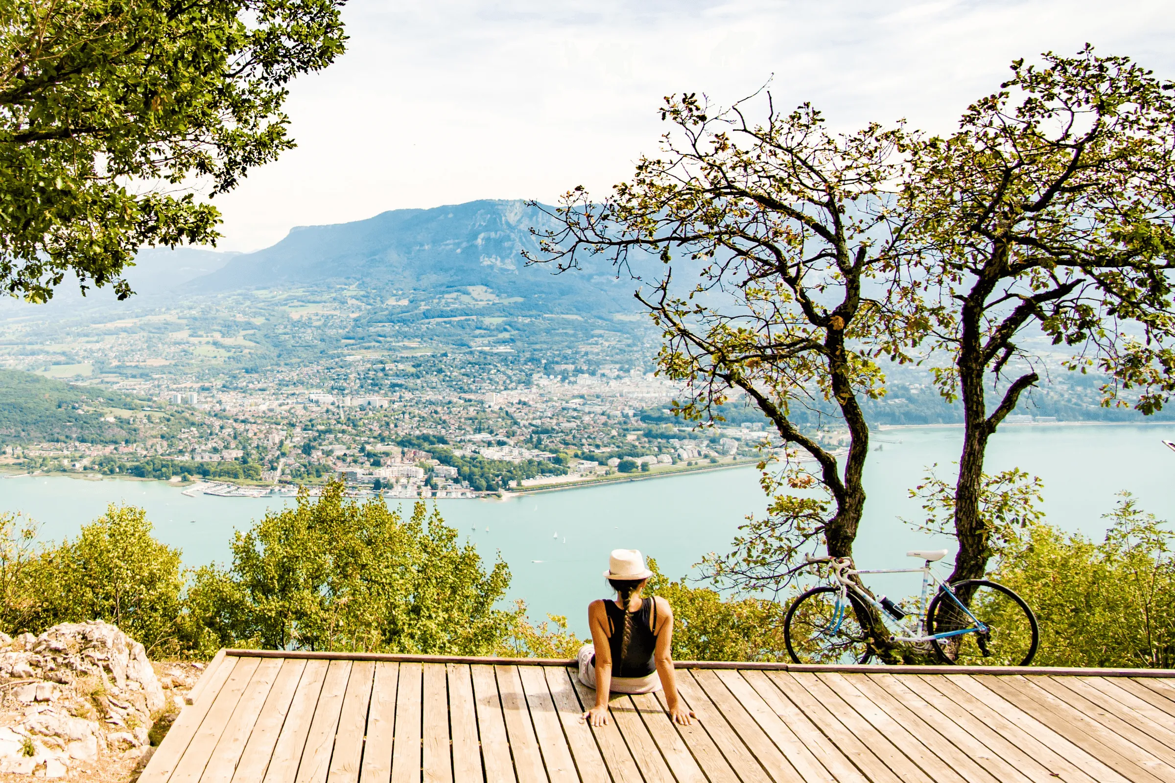 Une personne assise sur une plateforme en bois avec un vélo à côté, contemplant un lac et une ville entourée de montagnes