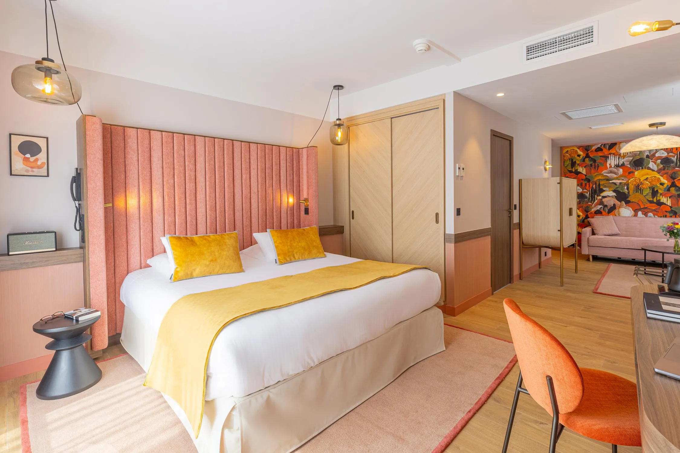 Chambre d'hôtel avec un lit double aux draps blancs et une couverture jaune, une tête de lit rose, une table de chevet avec des objets dessus, une lampe suspendue, un fauteuil orange et une porte ouverte sur un salon avec un canapé et une peinture murale