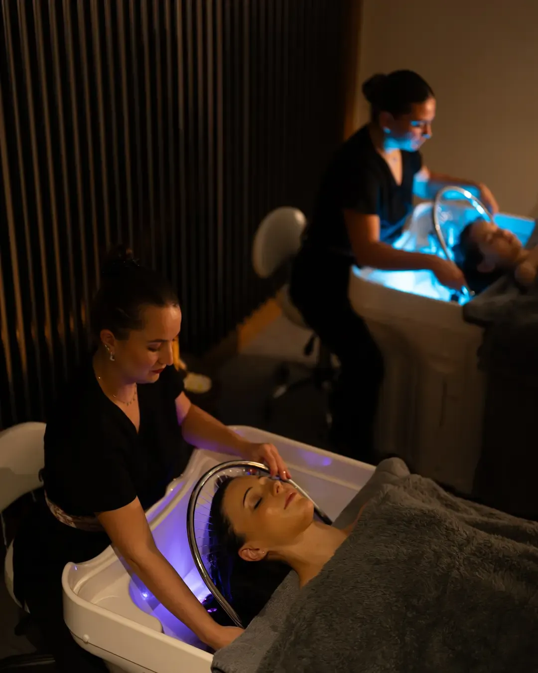 Two people receive a facial treatment in blue-lit devices, while two estheticians assist them in a room with dark curtains.